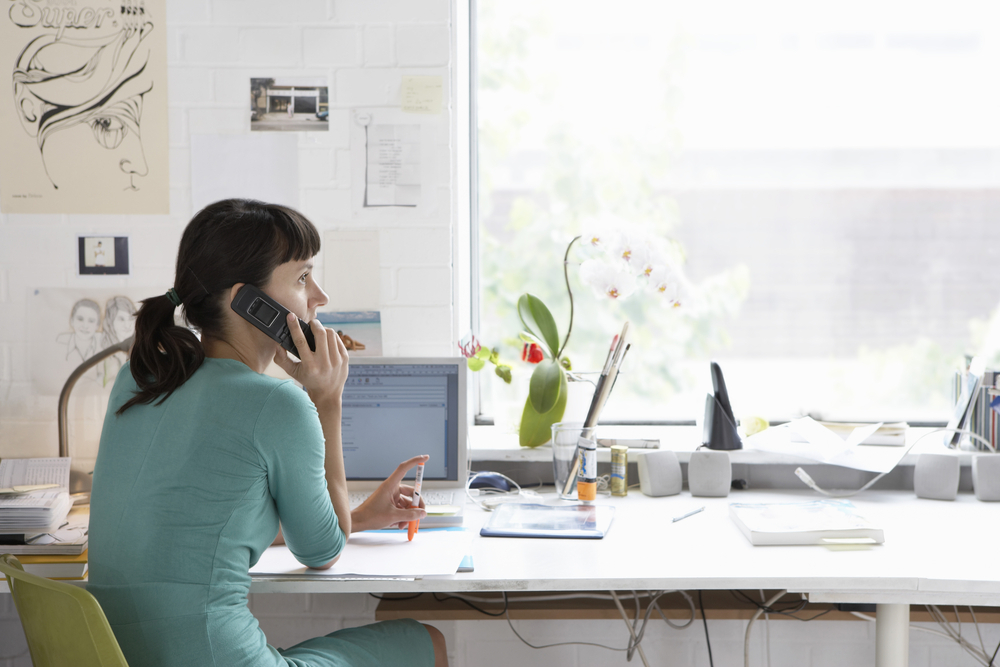 Mulher em seu escritório branco, com flores e alguns desenhos, conversando no telefone. 