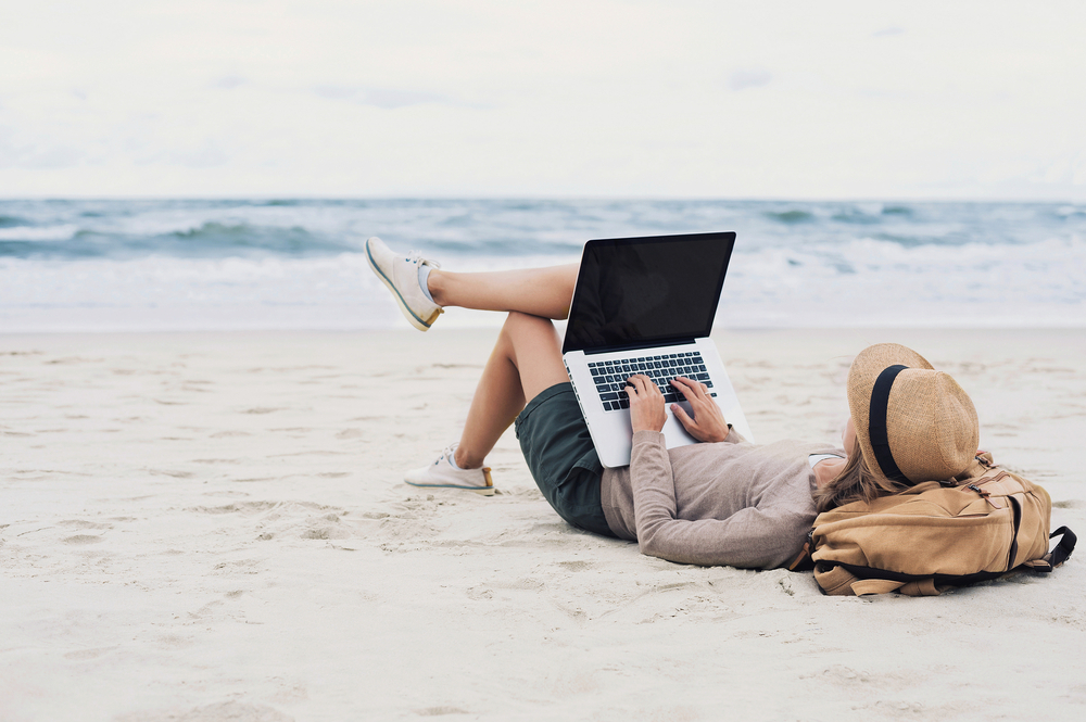 Mulher deitada na areia com roupas de verão, olhando para o mar e mexendo no computador